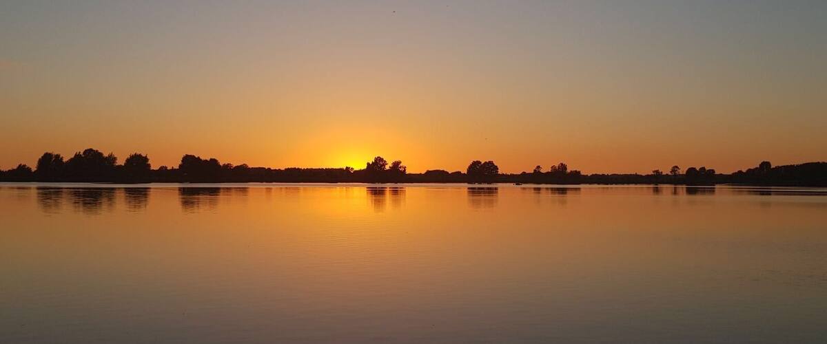 A calm lake at sunset with a clear sky and silhouettes of trees and a distant shoreline.