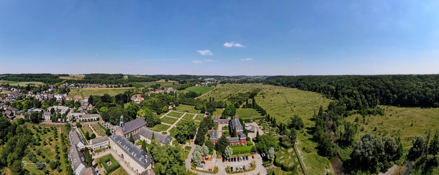 Aerial view of a monastery's imposing architecture nestled among manicured gardens and lush landscapes under a vast blue sky, Valkenburg, Limburg, Netherlands.