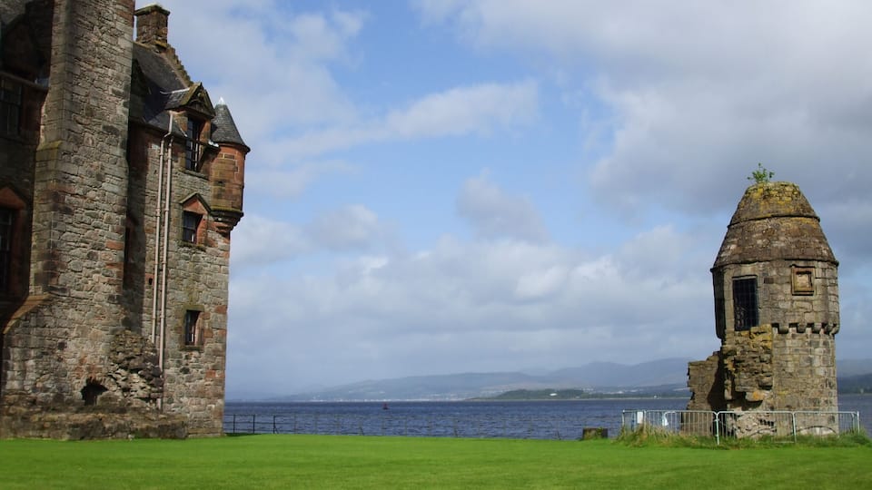 A historic stone building with a tower, a grassy field, and a body of water in the background.