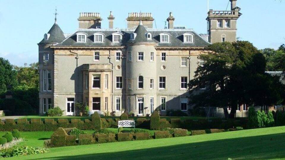 A large, historic building with a symmetrical facade, multiple chimneys, and a well-manicured lawn in front.