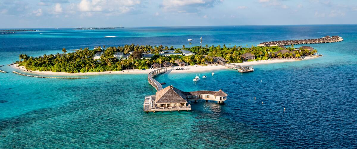 An aerial view of a tropical island resort with clear turquoise waters, palm trees, and overwater bungalows.