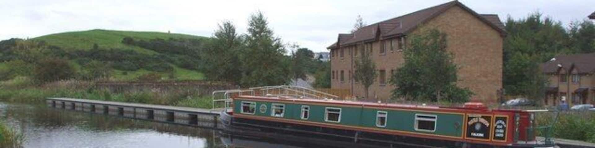 A narrowboat on a calm river, surrounded by greenery and a brick building.