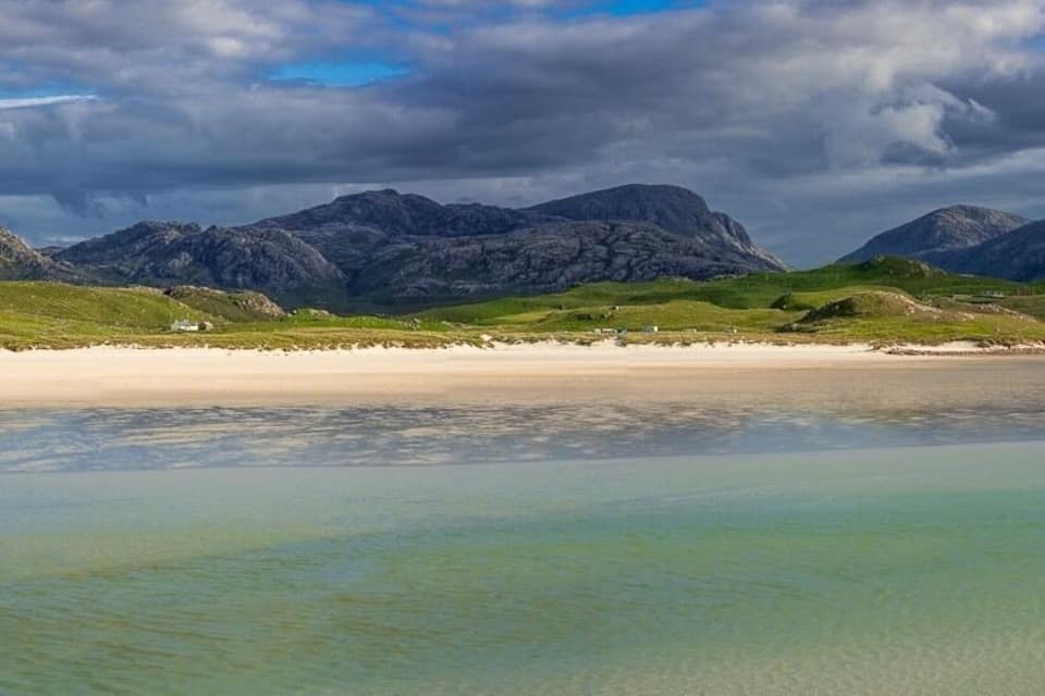 Un paysage côtier avec une plage de sable, des eaux turquoise et limpides, et des montagnes accidentées en arrière-plan.