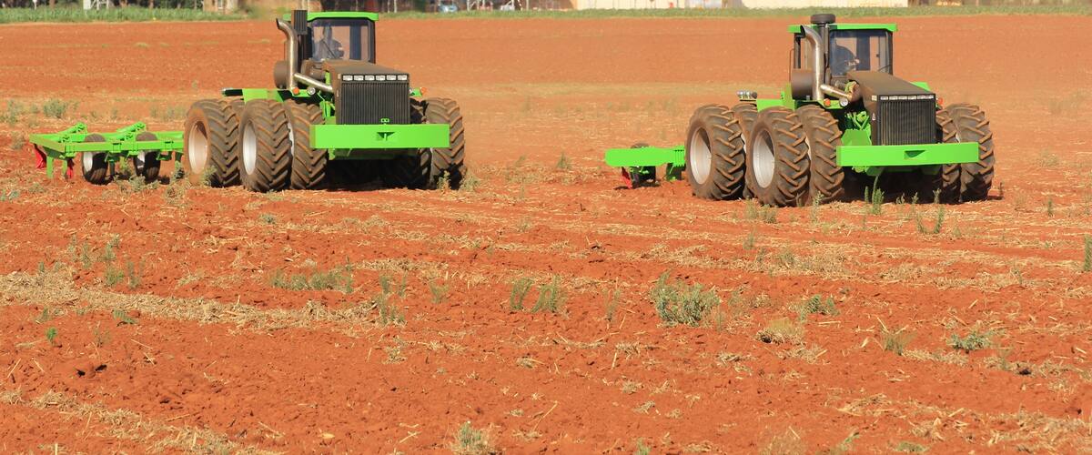 Two green tractors in a field with a red soil background and a white building in the distance.