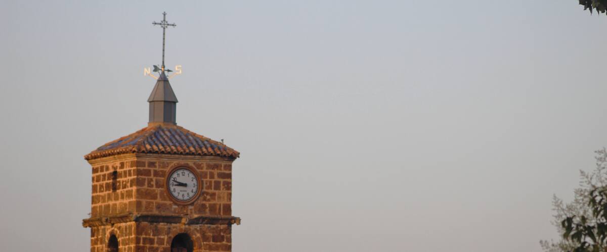 A stone clock tower with a cross on top, surrounded by buildings with red-tiled roofs.