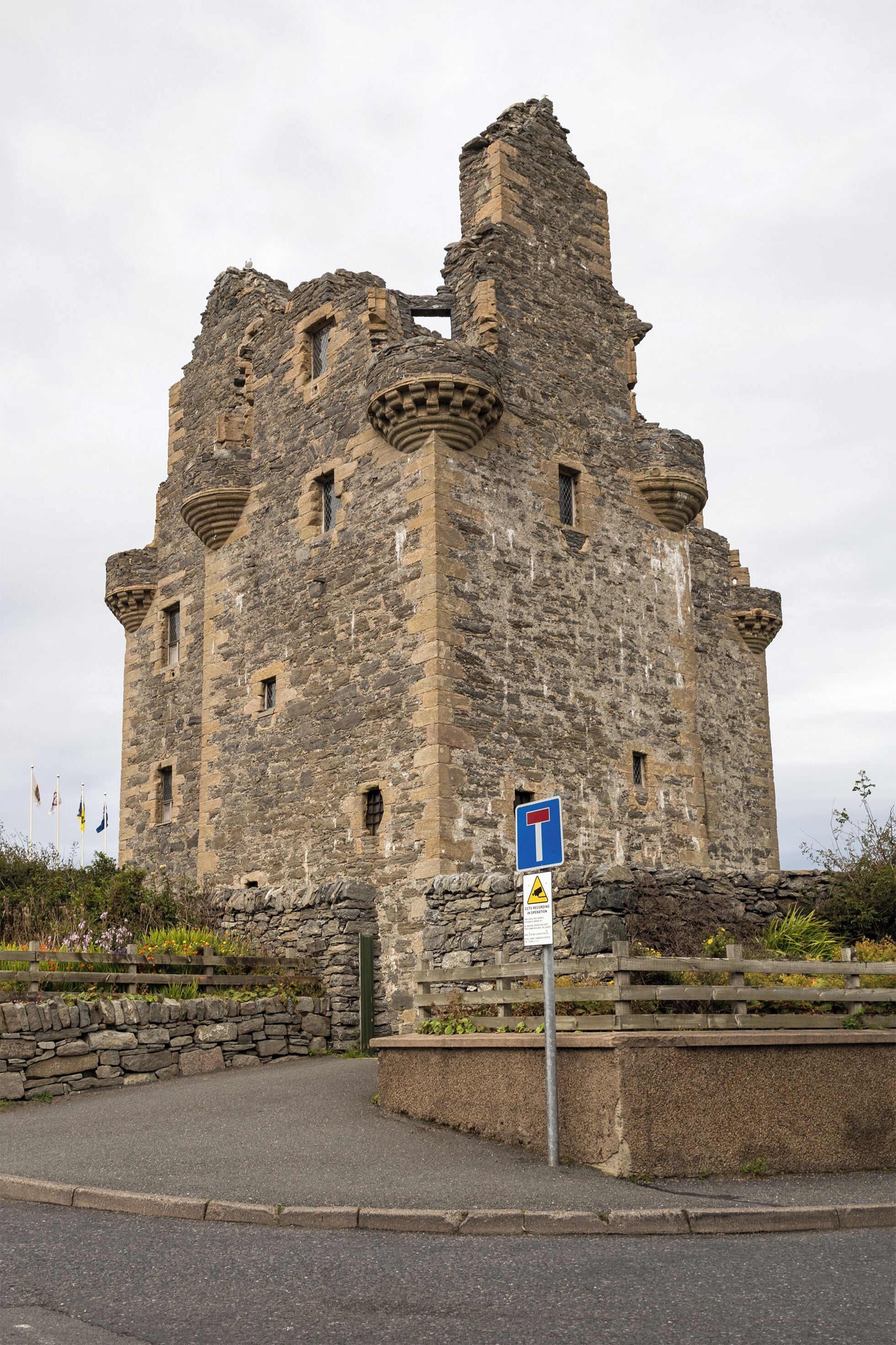 A historic stone castle with a round tower, a traffic sign, and a stone wall with vegetation.