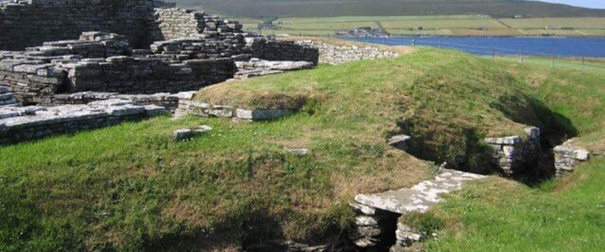 Des ruines antiques en pierre, situées au bord d’un lac, avec des collines verdoyantes en arrière-plan.