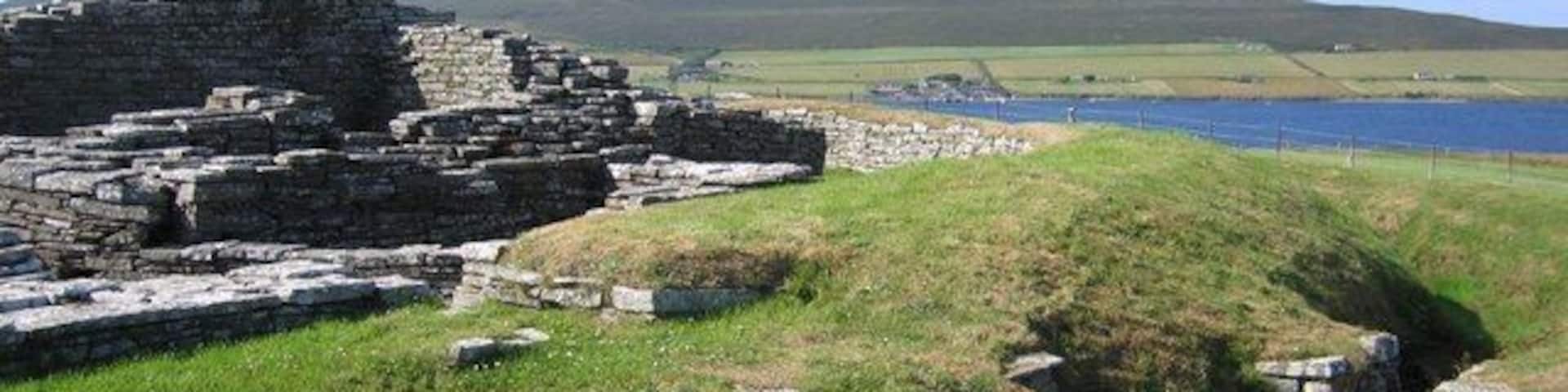 Ancient stone ruins by a lake with grassy hills in the background.