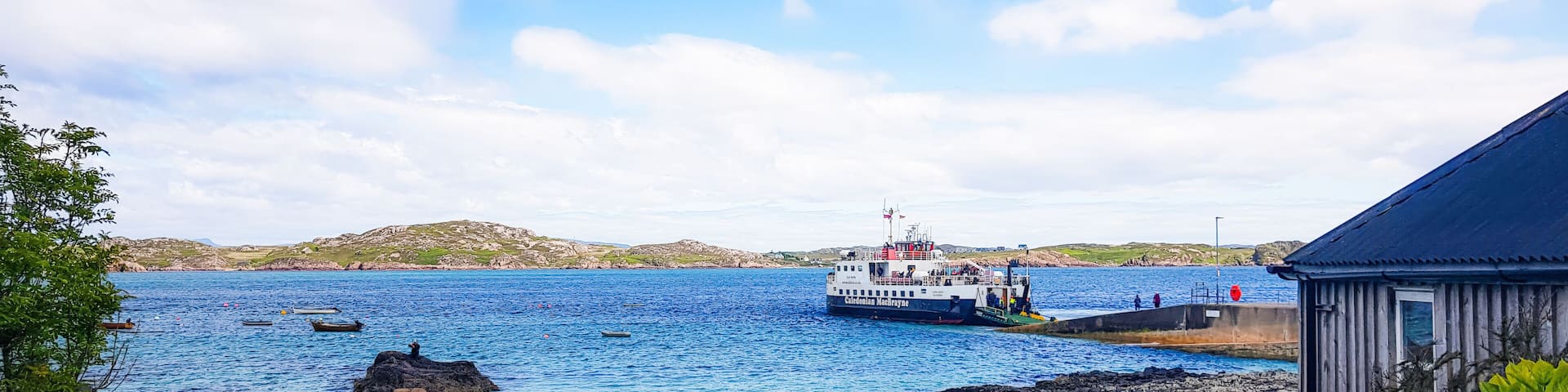 A coastal scene with a ferry docked at a pier, a red boat on the shore, and a small building.