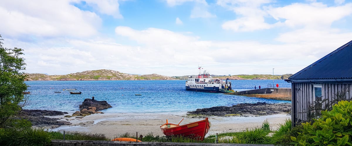 A coastal scene with a ferry docked at a pier, a red boat on the shore, and a small building.