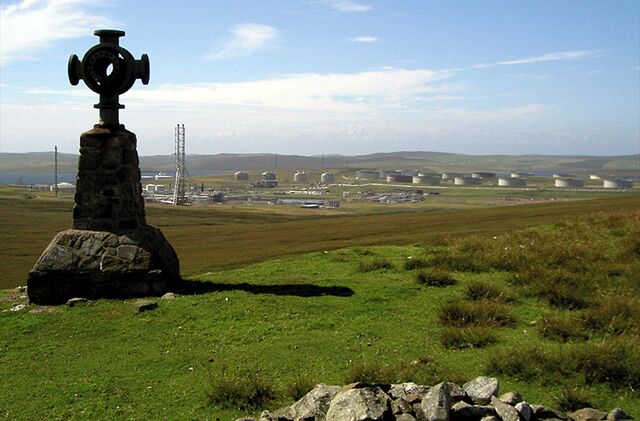 A stone monument with a circular top, a grassy field, and distant industrial buildings.