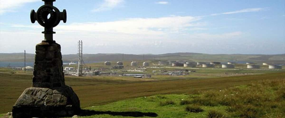 A stone monument with a circular top, a grassy field, and distant industrial buildings.