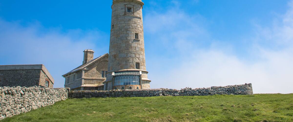 A stone lighthouse with a glass observatory at its base, surrounded by a stone wall and grassy hill.