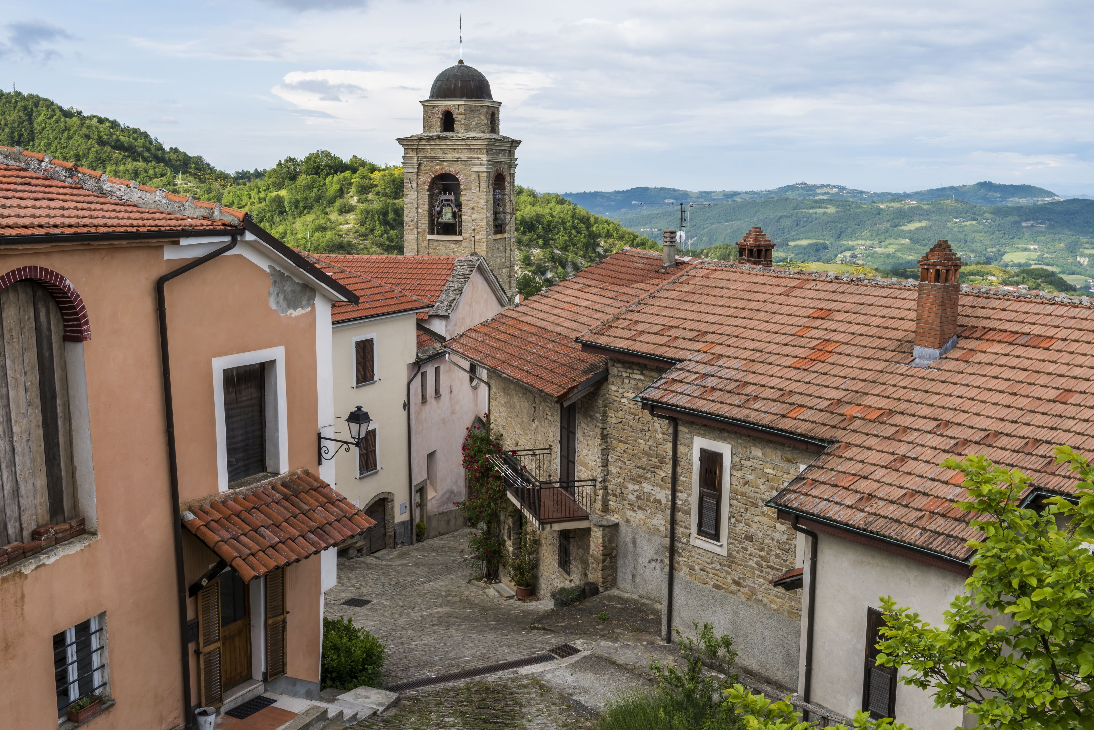 A narrow street with stone buildings, a bell tower, and a hillside in the background.