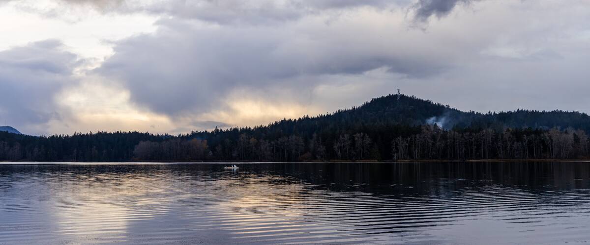 A calm lake with a forested hill in the background under a cloudy sky.