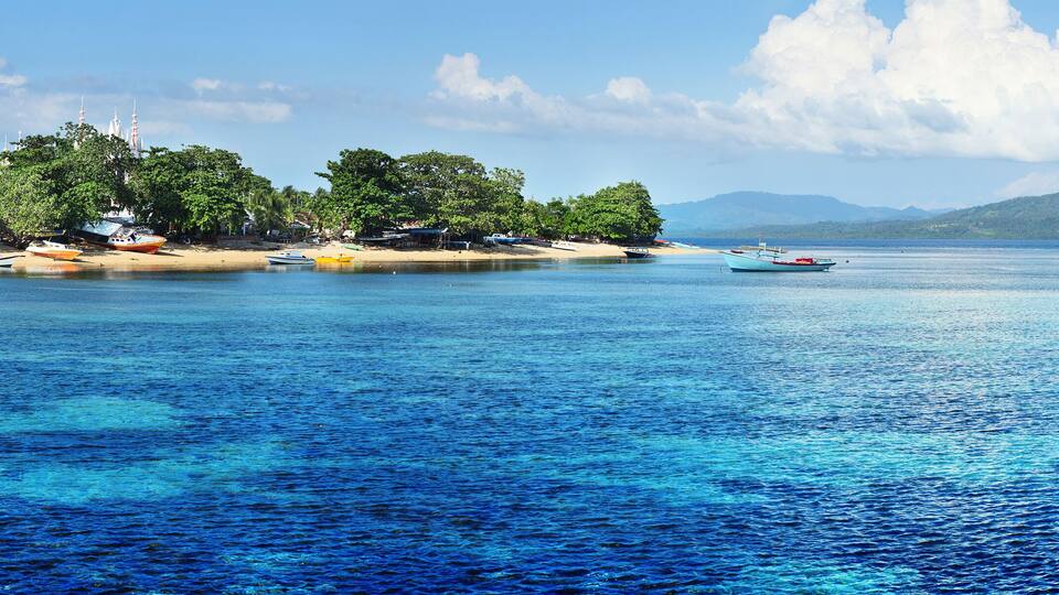 A tropical beach with clear blue waters, boats, and a mountain in the background.