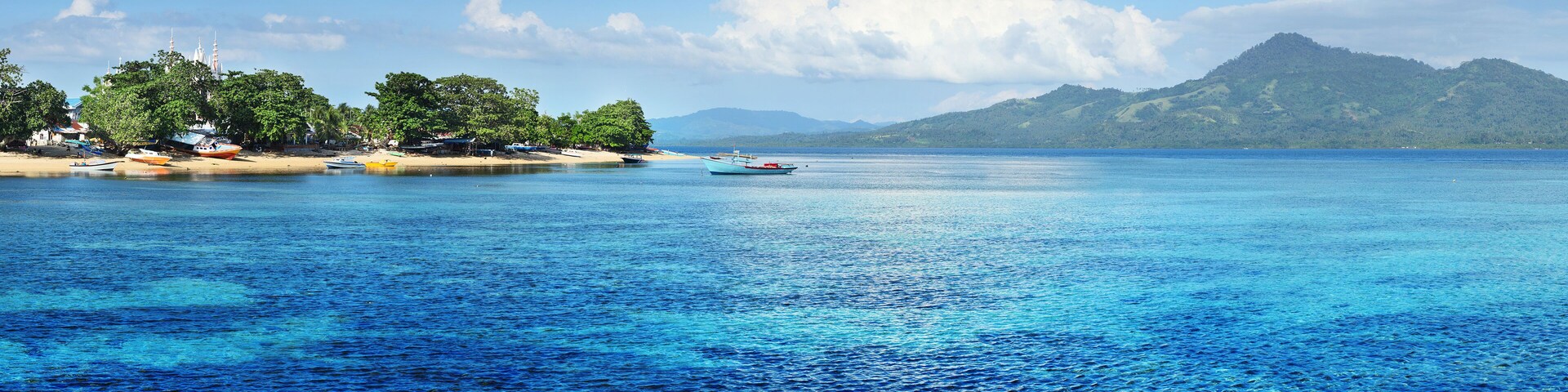 Uma praia tropical com águas azul-claras, barcos e uma montanha ao fundo.