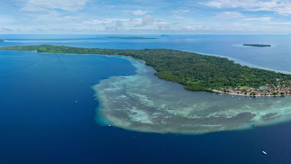 An aerial view of a tropical island with a crescent shape, surrounded by clear blue waters and a small town at its tip.
