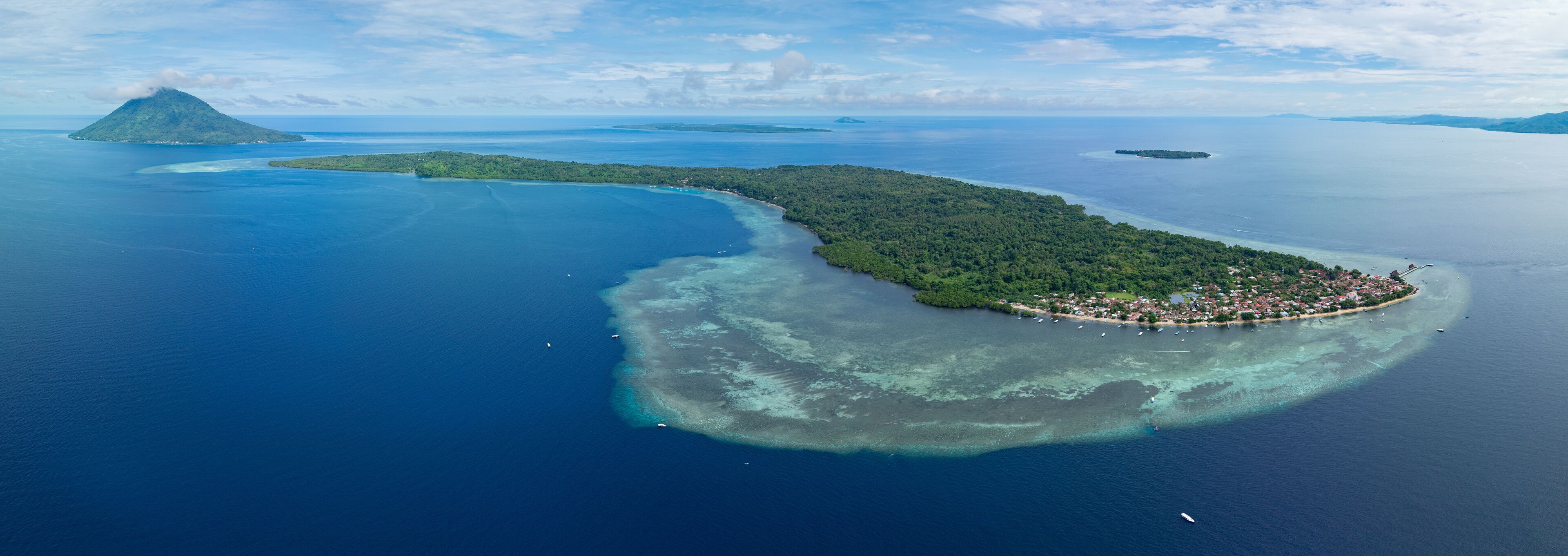 An aerial view of a tropical island with a crescent shape, surrounded by clear blue waters and a small town at its tip.
