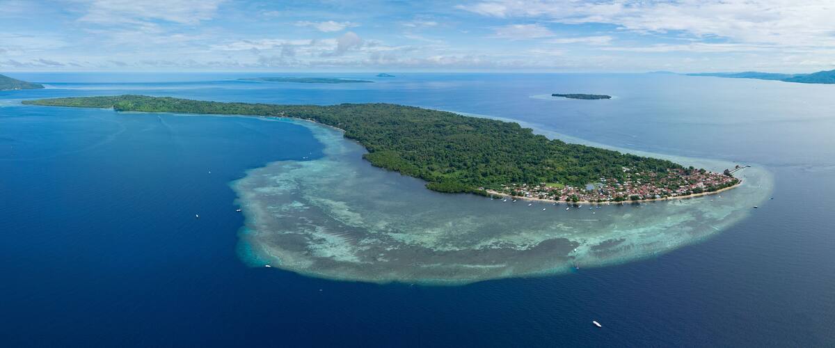 An aerial view of a tropical island with a crescent shape, surrounded by clear blue waters and a small town at its tip.