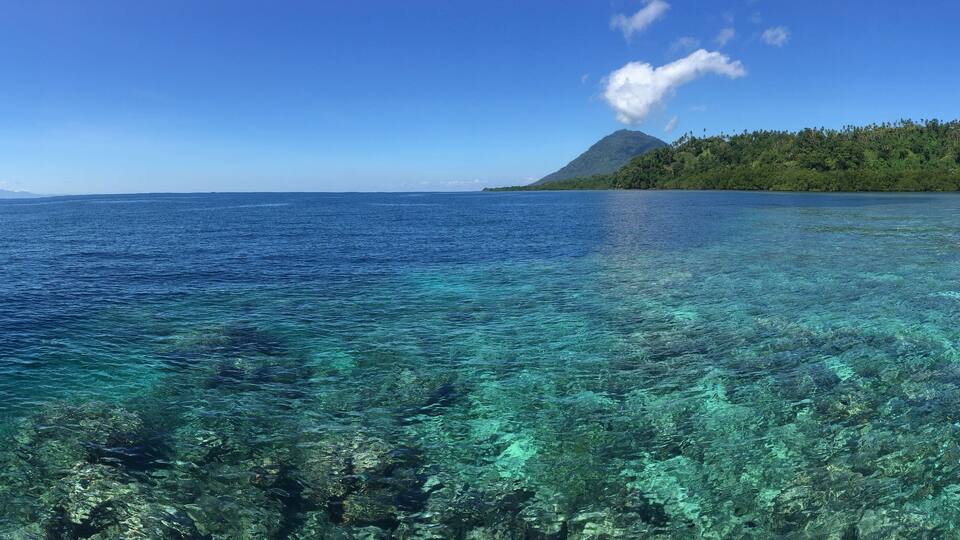 A clear blue sea with visible underwater coral reefs and a mountain with a smoke plume in the distance.