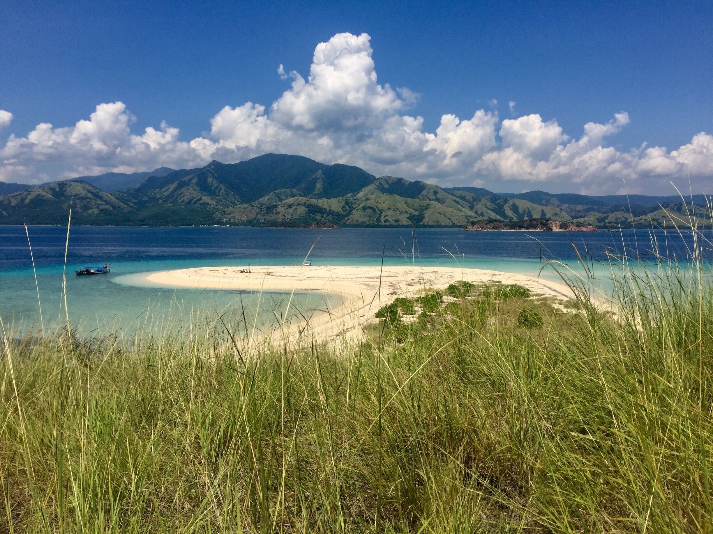 A beach with clear turquoise waters, a sandy shore, and green mountains in the background.