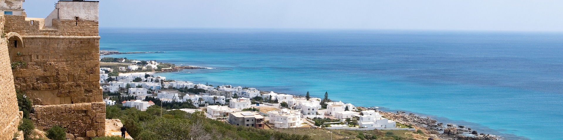 A coastal town with white buildings, a stone fort, and a clear blue sea.