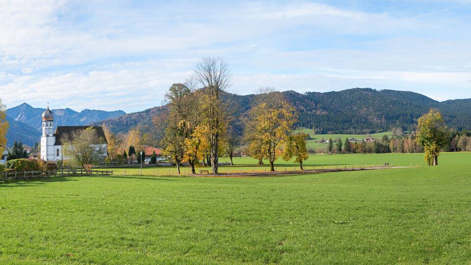 A rural landscape with a church, green fields, and autumn-colored trees.