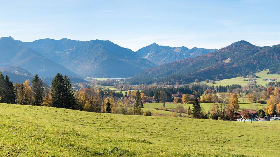 A mountainous landscape with a valley, green fields, and autumn-colored trees.