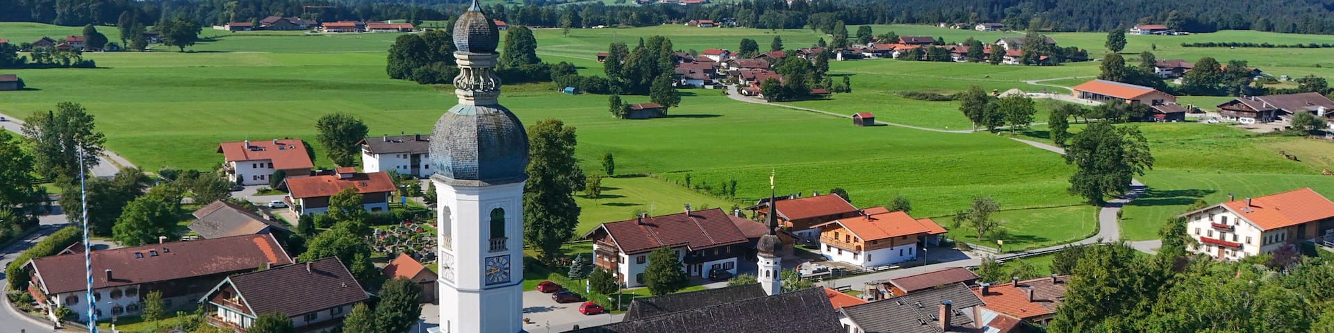 A town with a prominent church featuring a tall bell tower, surrounded by green fields and mountains in the background.