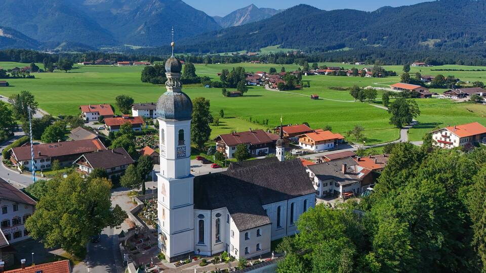 A town with a prominent church featuring a tall bell tower, surrounded by green fields and mountains in the background.