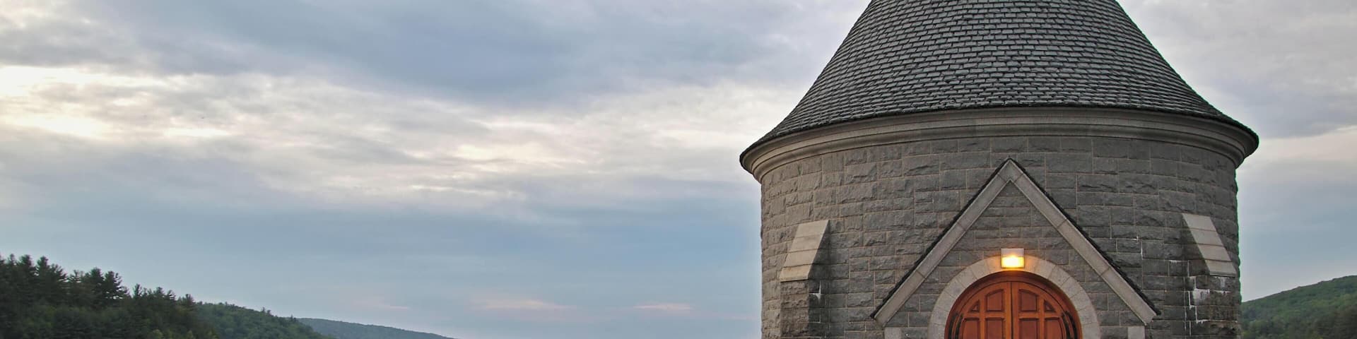 A stone tower with a conical roof by a calm lake, surrounded by forested hills.