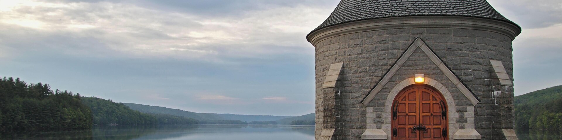 Uma torre de pedra com telhado cônico à beira de um lago calmo, rodeada por colinas arborizadas.