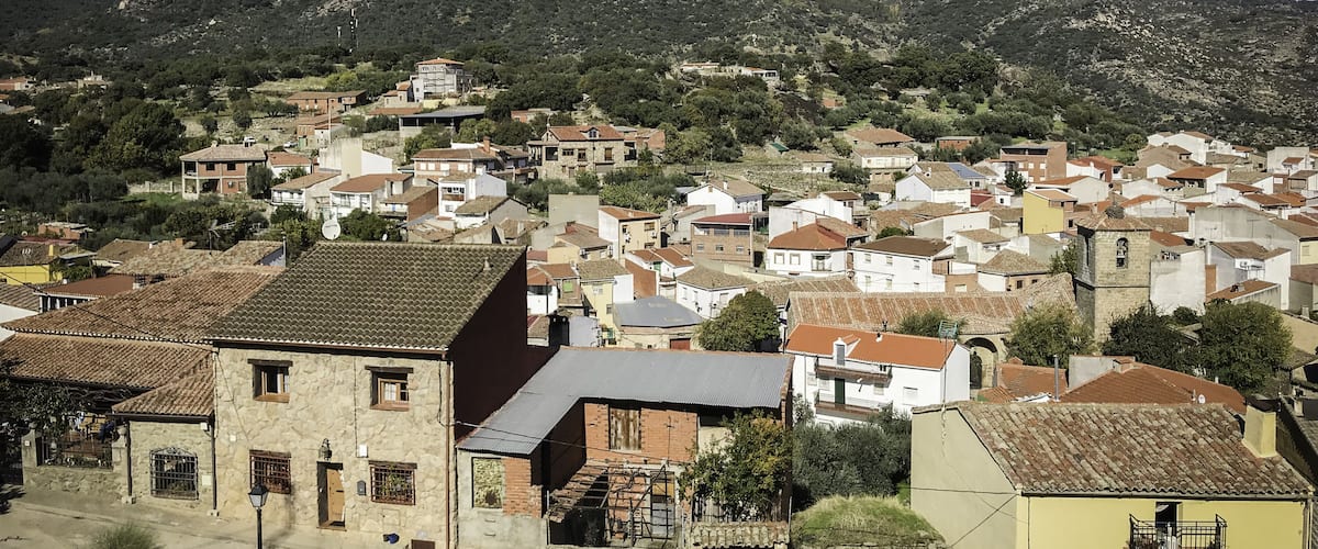 A village with terracotta-roofed houses, a prominent hill in the background, and a clear blue sky.