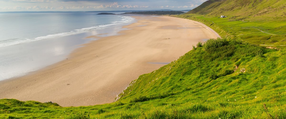 A coastal landscape with a sandy beach, grassy hills, and a distant white building.