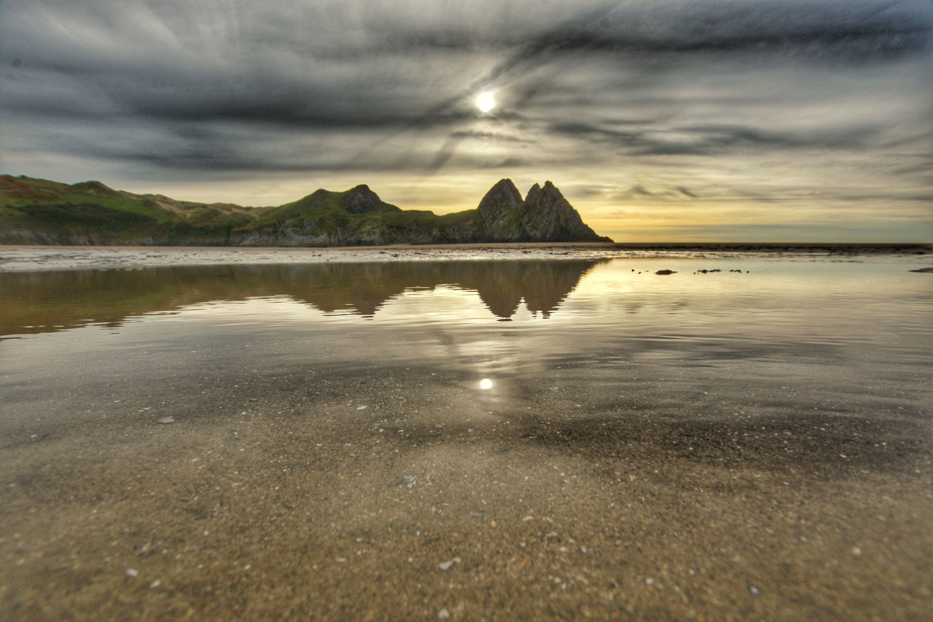 A beach with calm waters reflecting the sky and distant mountains.