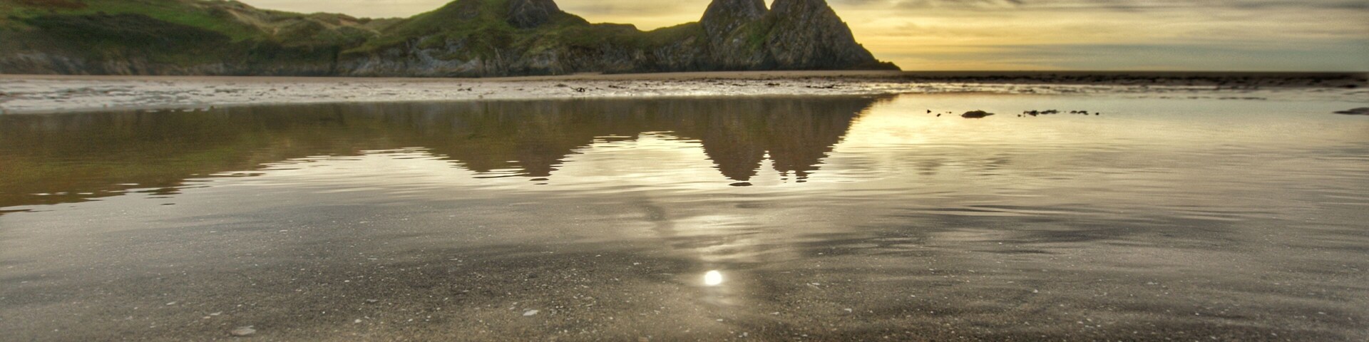 A beach with calm waters reflecting the sky and distant mountains.