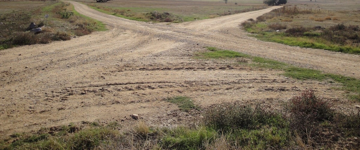 A dirt road in a rural landscape with sparse vegetation and distant hills.
