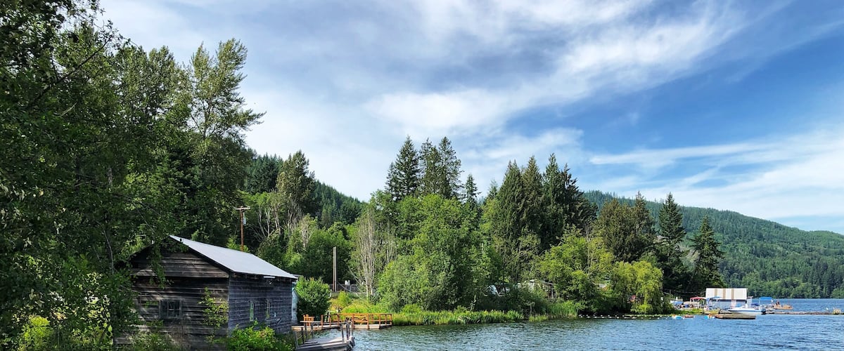 A wooden cabin by a calm lake surrounded by dense forest and hills.