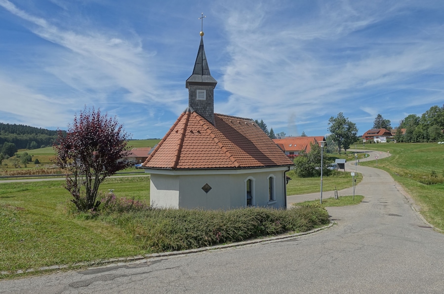 Una iglesia pequeña con tejado de tejas rojas y campanario, rodeada de campos verdes y un cielo azul despejado.