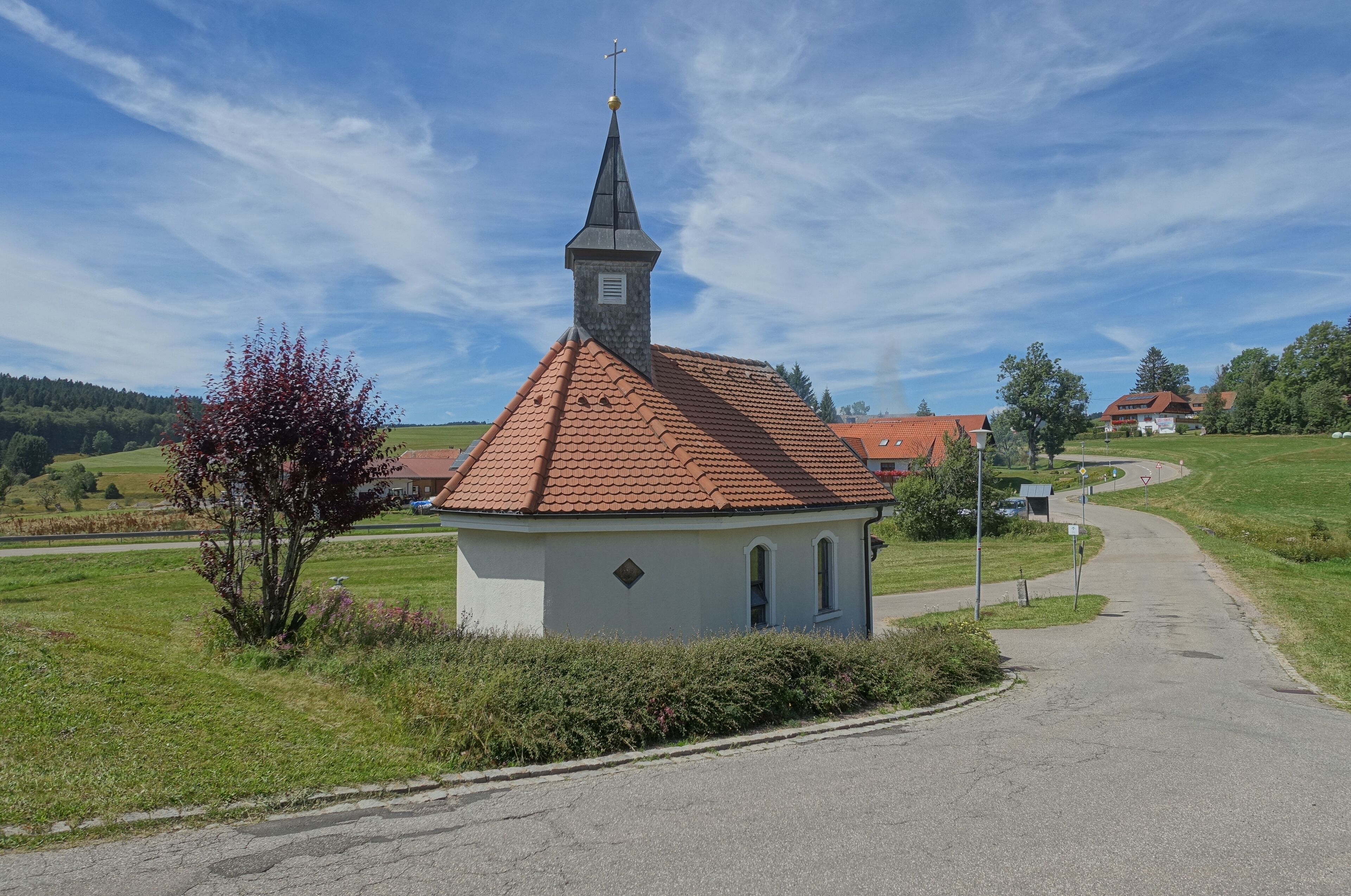 A small church with a red-tiled roof and a steeple, surrounded by green fields and a clear blue sky.