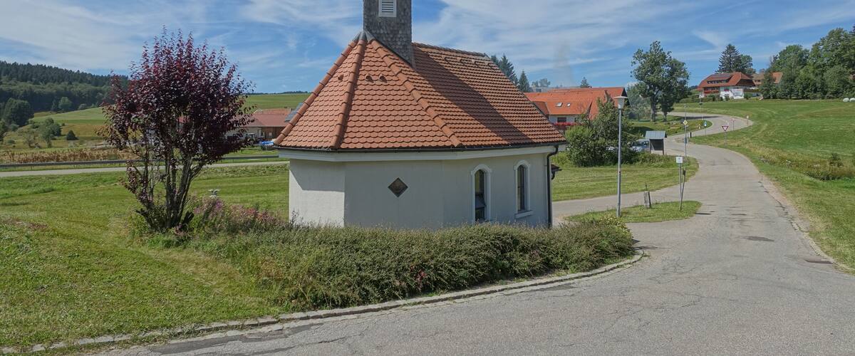 A small church with a red-tiled roof and a steeple, surrounded by green fields and a clear blue sky.