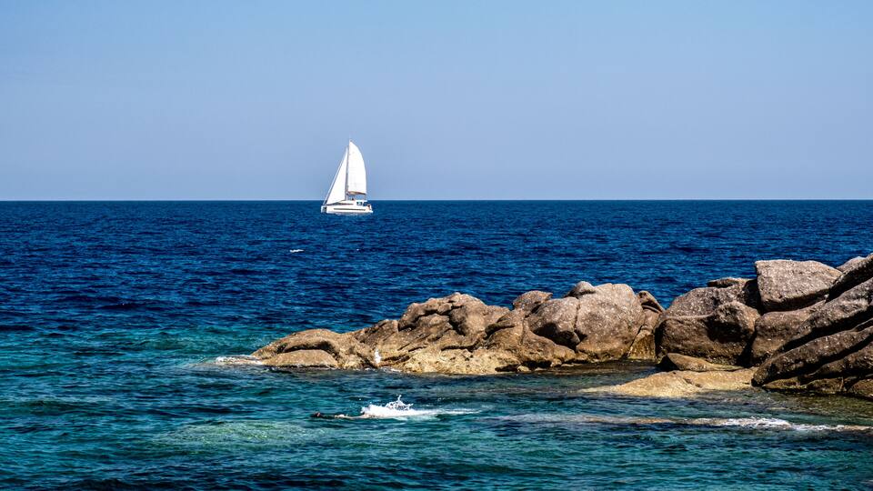 A sailboat on the ocean, rocky coastline, and clear blue water.