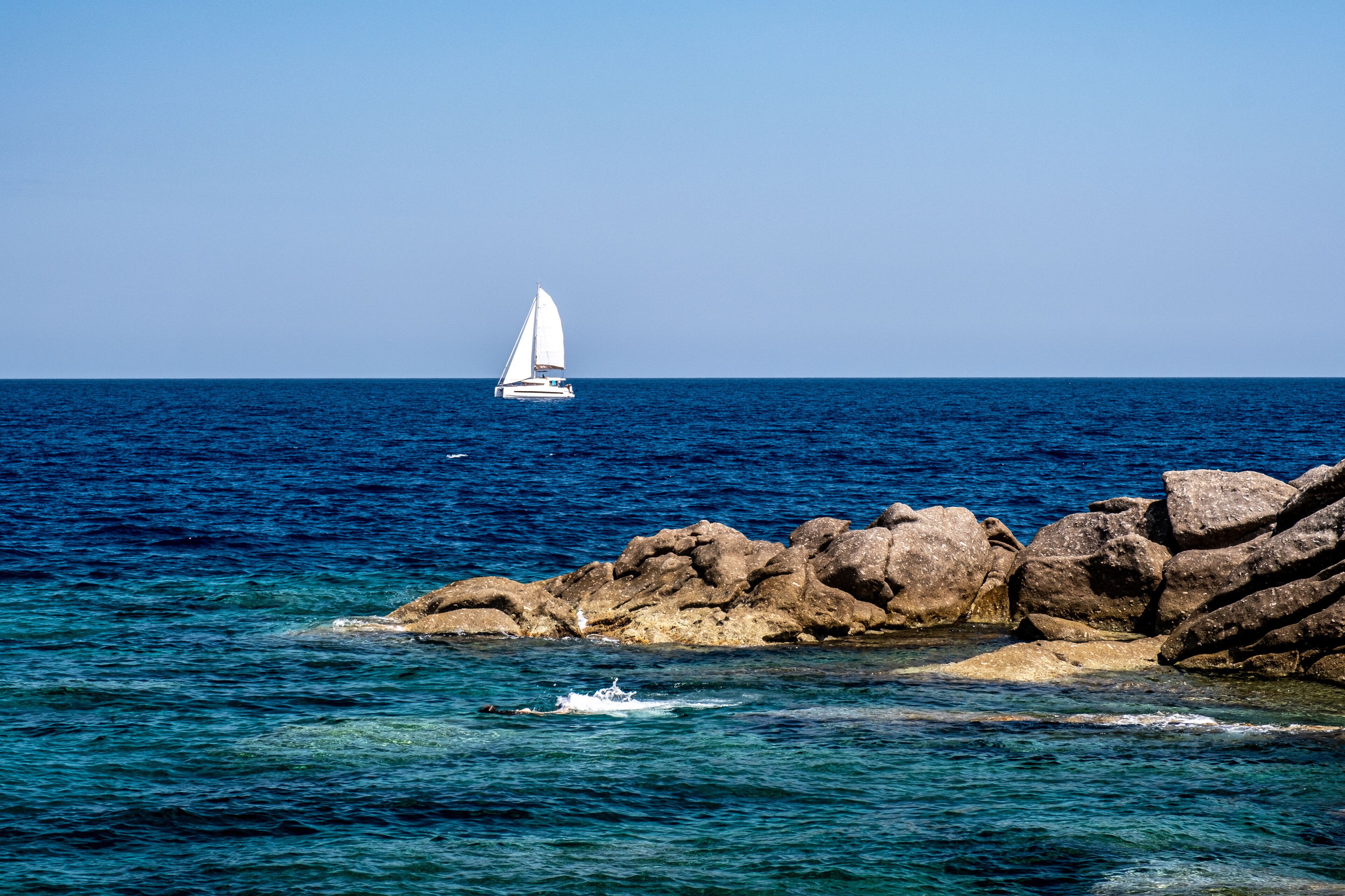 A sailboat on the ocean, rocky coastline, and clear blue water.