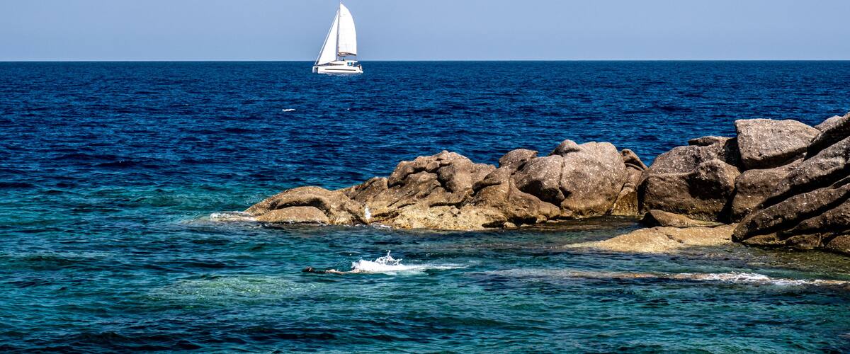 A sailboat on the ocean, rocky coastline, and clear blue water.
