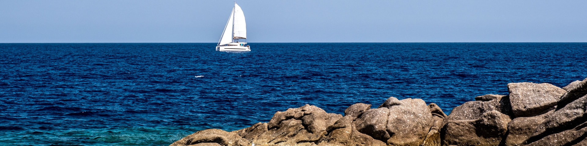 A sailboat on the ocean, rocky coastline, and clear blue water.