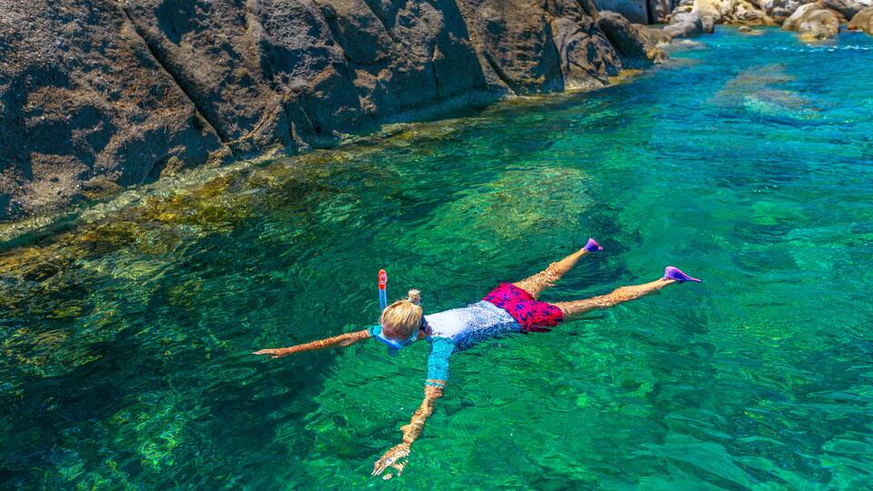 A person snorkeling in clear blue water with rocky formations in the background.