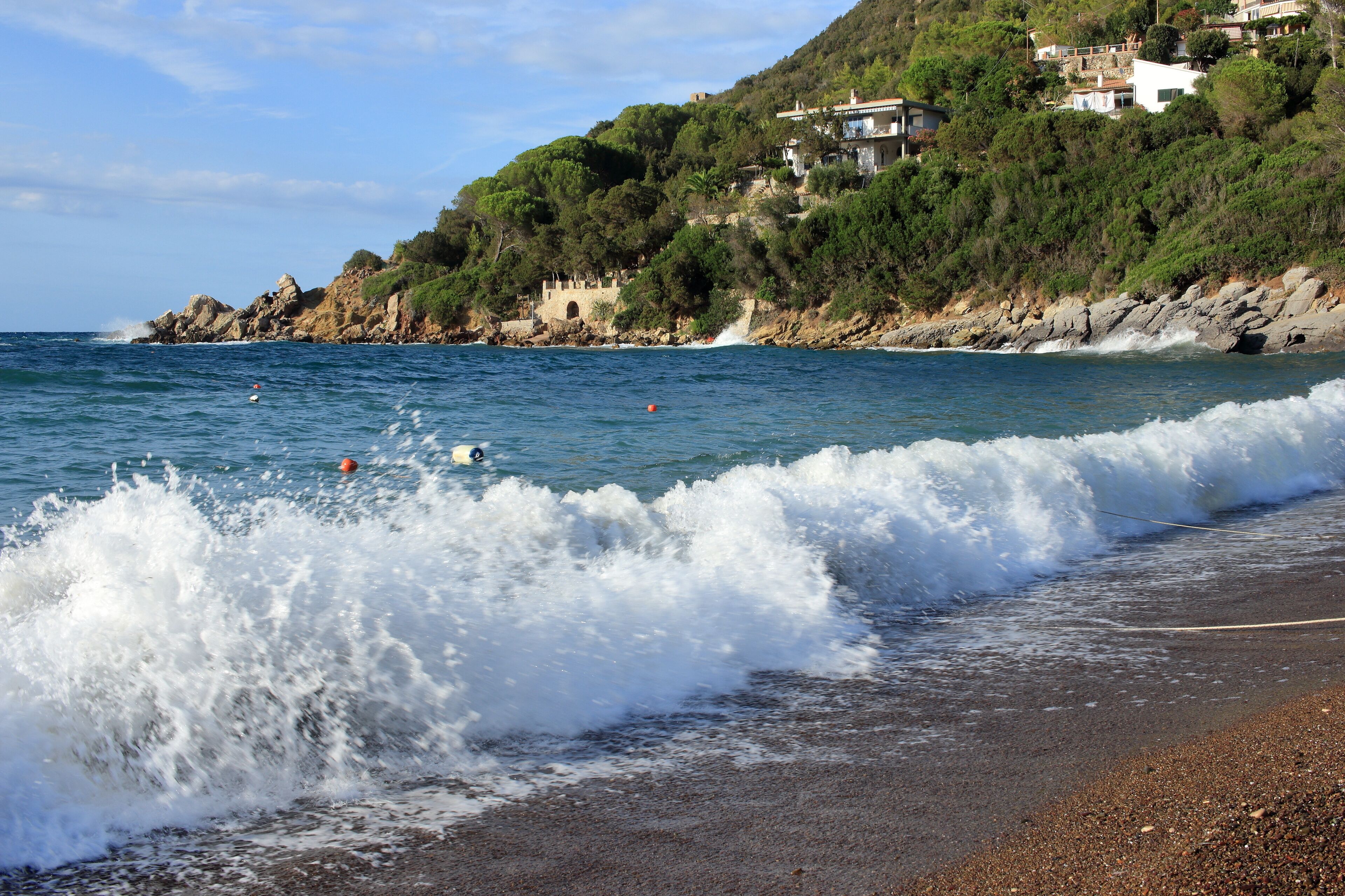 A beach with waves crashing onto the shore, a rocky coastline, and houses on a hillside.