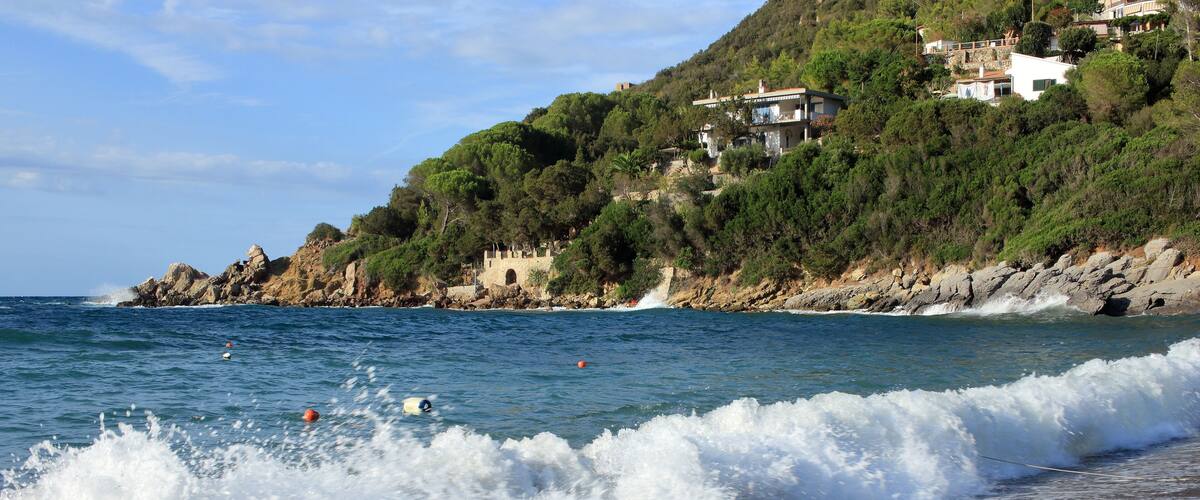 A beach with waves crashing onto the shore, a rocky coastline, and houses on a hillside.