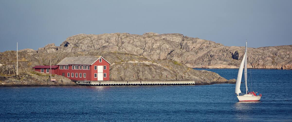 A red house by a rocky shore with a sailboat in the water.